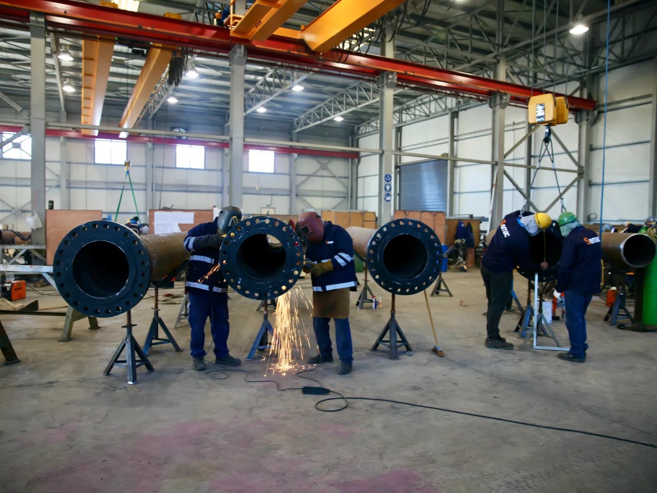 Welders working on large pipe sections on stands in the workshop, under an overhead crane