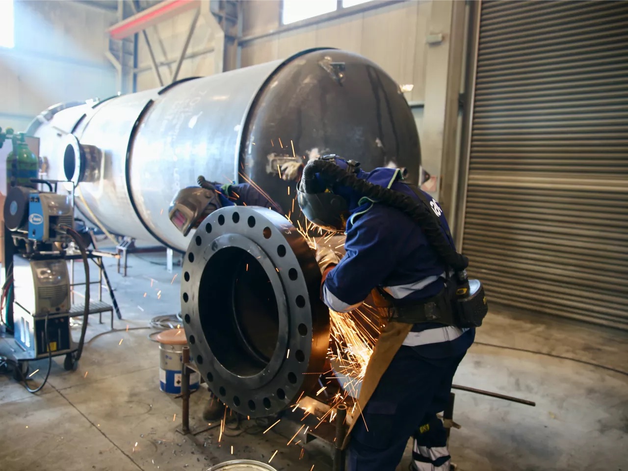 A technician with a grinder at the flange of a large pipe section in the workshop