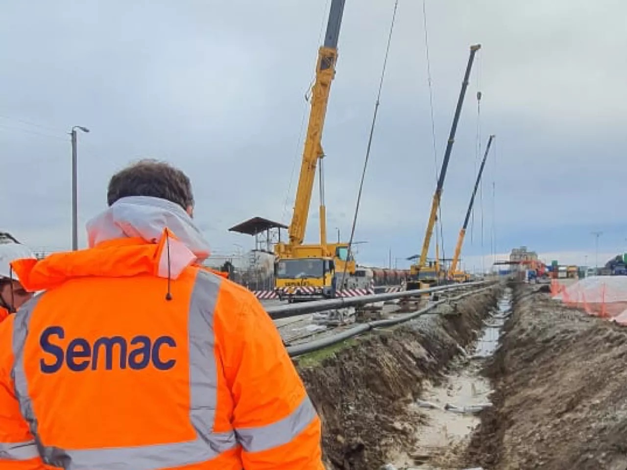 A SEMAC team member in a high-visibility jacket at a pipeline site, with cranes and an open trench